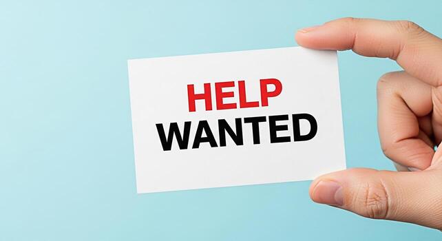 Hand holding a white card with the text Help Wanted in red and black font against a light blue background symbolizing job openings recruitment and the need for assistance in a business or organization photo