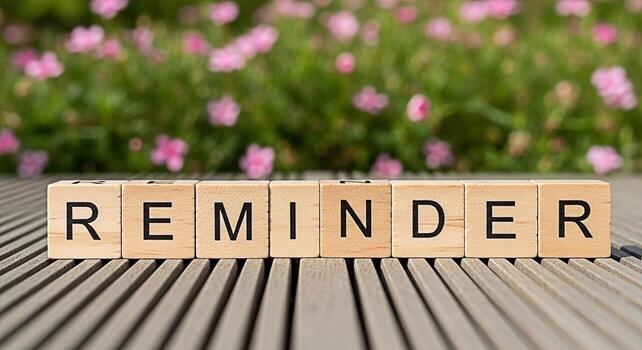 Wooden blocks spelling REMINDER placed on a wooden surface with a blurred background of pink flowers and green foliage creating a gentle and encouraging atmosphere for important tasks and deadlines photo