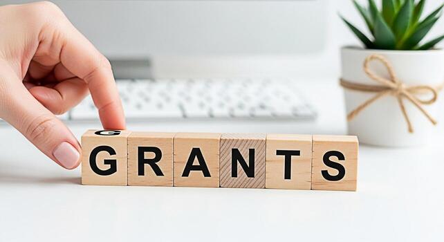 Female hand arranging wooden blocks spelling GRANTS on a white desk with a keyboard and a succulent symbolizing funding opportunities and financial support for projects and initiatives in a clean mode photo