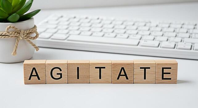 Wooden blocks spelling Agitate on a white desk with a keyboard and succulent representing the concept of stirring up emotions debate or action in a clean modern workspace conveying a sense of urgency photo
