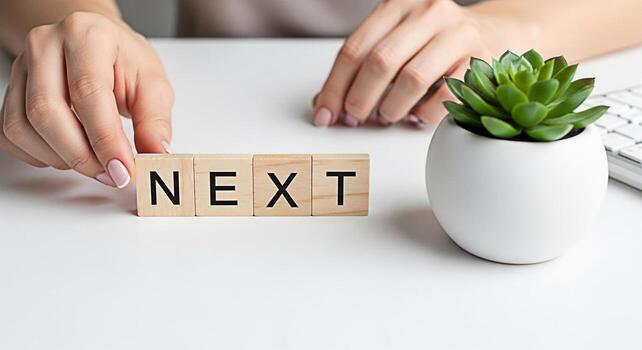 Womans hand arranging wooden blocks spelling NEXT on a clean white desk next to a succulent symbolizing anticipation and forwardthinking in a bright minimalist workspace conveying a sense of progress photo