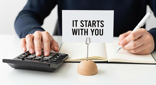 Focused professional calculating finances and writing in a notebook at a bright desk emphasizing personal responsibility and motivation with a card stating It Starts With You for financial planning an photo