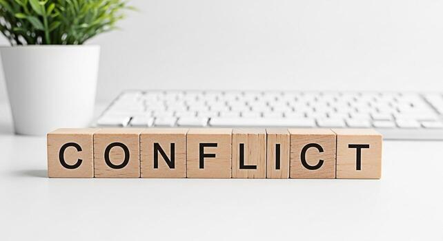 Wooden blocks spelling CONFLICT on a white desk with a keyboard and potted plant in the background representing disagreement tension and the need for resolution in a modern workplace environment photo