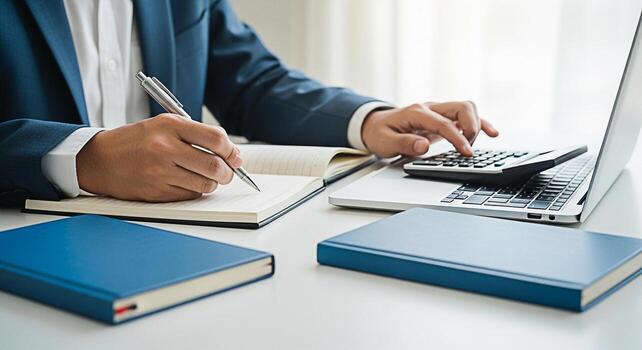 Diligent businessman in a bright office calculates finances using a calculator on a laptop while taking notes in a notebook conveying a sense of focus and financial planning for business success photo