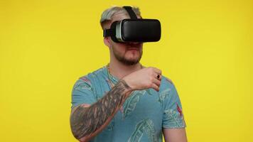 förvånad turist vuxen man använder sig av headsetet hjälm app till spela simulering spel. tittar på virtuell verklighet 3d 360 . ung kille i vr glasögon isolerat på gul bakgrund. missbruk från ny teknologi video