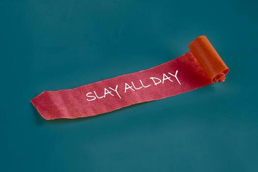 A red textured paper with white handwriting that says SLAY ALL DAY is partially unrolled against a Teal background in a studio setting, promoting confidence and goals photo