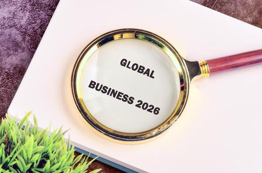 Magnifying glass focuses on text reading GLOBAL BUSINESS 2026. A white notepad sits beneath with artificial greenery in the corner. A symbol of looking forward photo