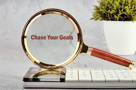 A magnifying glass rests on a keyboard, highlighting the text Chase Your Goals. A potted plant is in the background, signifying growth and achievement in business photo