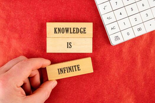 Business, limited or unlimited concept. Words KNOWLEDGE IS INFINITE assembled from blocks in a composition with part of a white calculator and a hand, top view photo