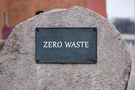 The words ZERO WASTE are displayed on a dark rectangular plaque affixed to a large, textured grey stone. It suggests a commitment to reduce pollution photo