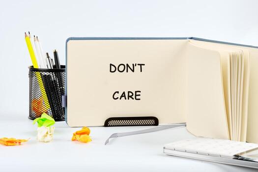 The scene contains an open notebook displaying the words Dont Care. Nearby are writing utensils in a metal container, plus crumpled paper, and a notebook, all on a white surface photo