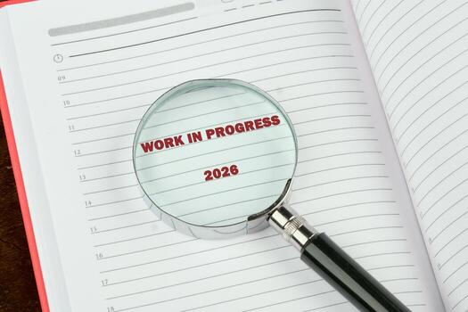A lined notebook, opened to a blank page, is viewed through a magnifying glass. The words WORK IN PROGRESS 2026 are highlighted by the magnifying lens photo