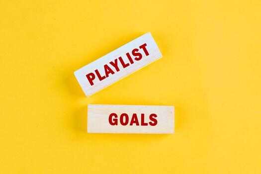 On a bright yellow background, two wooden blocks are placed showing the words PLAYLIST and GOALS, suggesting the act of creating musical experiences with intentional song choices photo