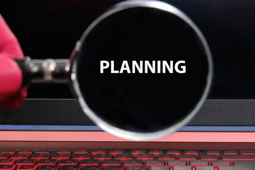 A person uses a magnifying glass to clearly view the word planning on a laptop screen. The glowing keys and dark background create a focused workspace atmosphere photo