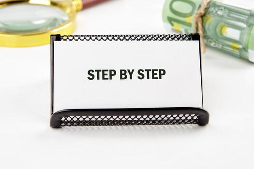 A small black stand holds a white card that reads STEP BY STEP, surrounded by a magnifying glass and rolled currency. The setting suggests a focused approach to tasks photo