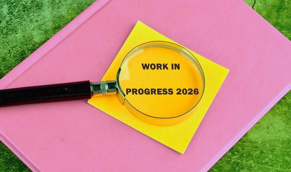 A magnifying glass is positioned over a bright yellow sticky note that reads work in progress. The note is placed on a soft pink notebook, all set against a vibrant green background photo