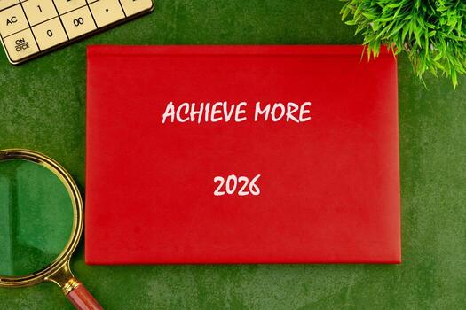 The bright red notebook with Achieve More on its cover sits on a green table, inviting creativity and ambition. A magnifying glass and a plant add thoughtful details to the workspace photo