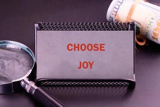 A sleek business card holder displays the message Choose Joy, surrounded by a magnifying glass and rolled money, blending motivation with finance in a modern workspace setting photo