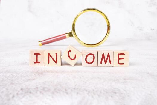 Blocks spelling out income sit on a soft surface, while a magnifying glass emphasizes the word, inviting viewers to explore financial concepts and opportunities in depth photo