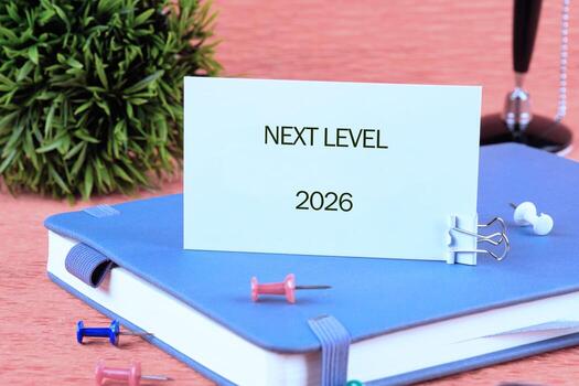 A workspace is set up with a blue notebook, colorful push pins, and a small green plant, displaying a card that reads next level, highlighting future goals for 2026 photo