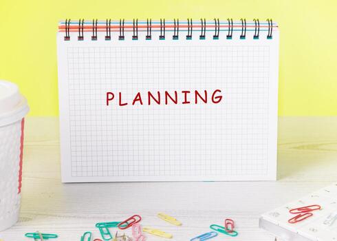 A notebook with the word planning in bold red letters sits on a light wooden surface surrounded by paper clips and a coffee cup, creating a vibrant workspace atmosphere photo
