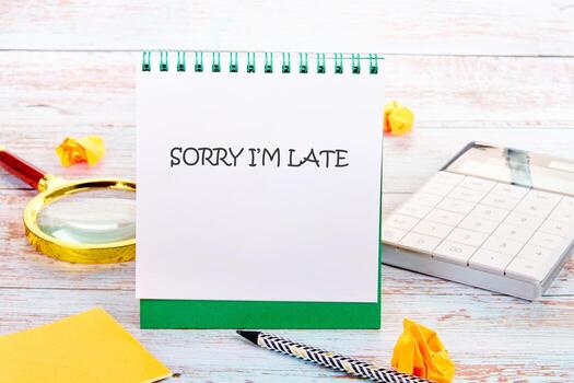 A desk displays a spiral notepad reading Sorry Im Late. A calculator, pen, magnifying glass, sticky note, and crumpled paper add to the scene, suggesting a busy day photo