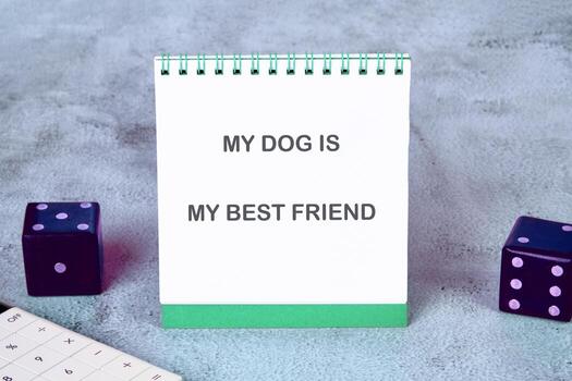 Close up of a notepad stating My Dog is My Best Friend. Dice sit on either side of the notepad, and a calculator is partially visible. All are sitting on a mottled grey surface photo