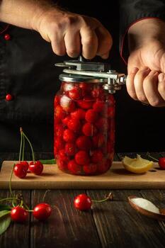 The chef rolls up a jar with a cherry drink or compote with a manual seaming key in his hands. The concept of preserving cherries in jars for future use. Advertising space on the black table photo