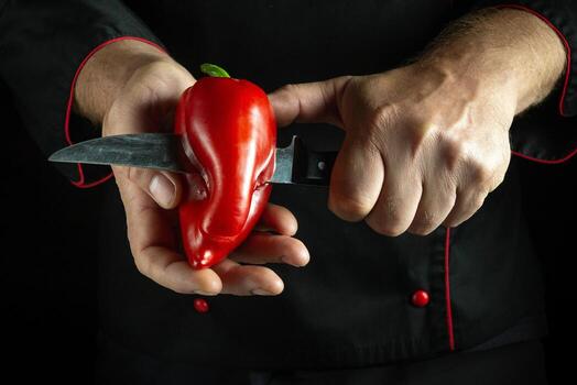 With deft hands, a chef holds a vibrant red pepper while expertly slicing it with a sharp knife, showcasing culinary precision in a dramatic, low-light setting photo