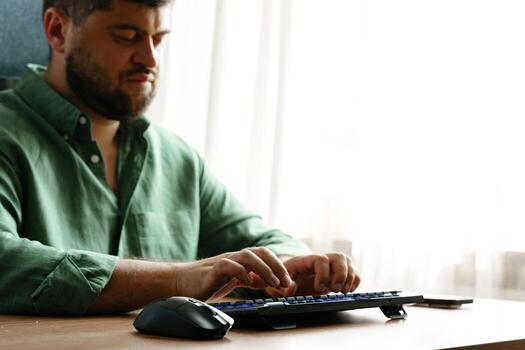 Man typing on keyboard while sitting at desk by window in a well-lit room photo