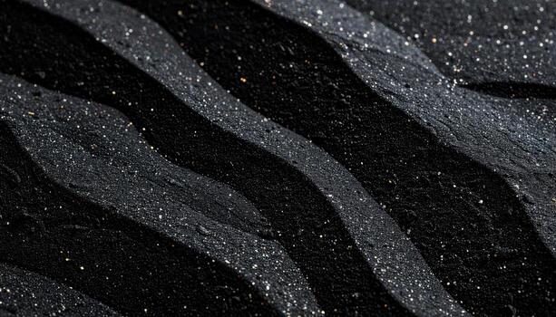 Black sand ripples glisten with light reflecting off of tiny grains in a close-up, textured, abstract pattern on a natural beach surface. photo