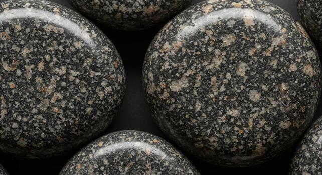 Close-up showcases smooth, round dark granite stones with speckled beige and gray inclusions on a matte black surface, creating a natural pattern. photo