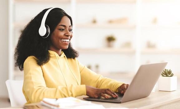Intelligent millennial black woman attending online class from home, sitting at table in front of laptop, taking notes, using headset, typing on computer keyboard, copy space. E-education concept photo