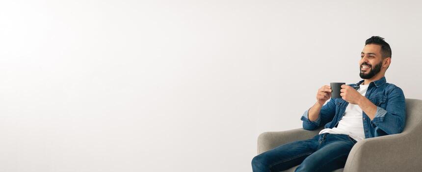 A man sits relaxed in a soft chair, sipping coffee with a smile. The bright room has a simple design, creating a calm and inviting atmosphere. He enjoys his moment of peace and joy. photo