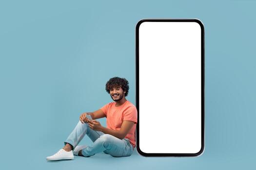 Curly-haired Indian man sits casually next to a giant smartphone with a blank screen. He enjoys using his phone while smiling, creating a relaxed and friendly atmosphere in a blue studio. photo