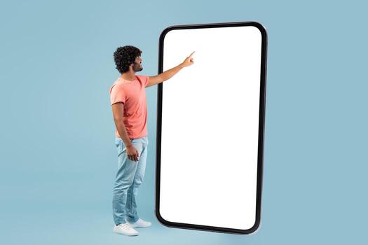 A fashionable young hindu man in casual attire is touching a large empty smartphone screen. He is focusing on the device with a blue studio background, exploring new mobile app options. photo