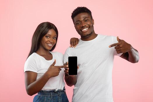 A young black couple stands in front of a pink wall, smiling and pointing at a blank smartphone screen. They are excited to share a new application, creating a friendly atmosphere for the viewer. photo