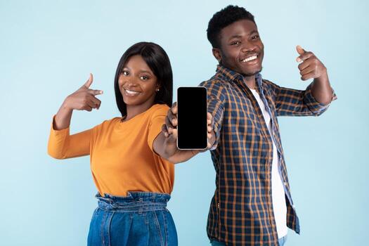 Black couple is smiling and pointing at a blank smartphone screen, presenting it for a mockup. They stand in a blue studio background, promoting a new mobile app together. photo