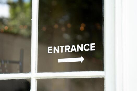 White entrance text and arrow are displayed on a clear glass door, indicating direction for entry. Suitable for use in wayfinding, navigation, or business signage contexts. photo