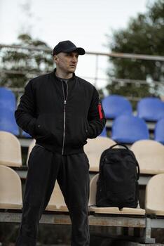 Man in black bomber jacket and cap standing alone in empty stadium tribune with backpack during cloudy day photo