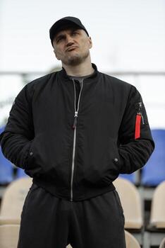 Man in black bomber jacket and cap reacts in empty stadium during intense game scenario photo