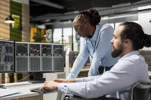 Risk analyst and coworker with disability in inclusive office using trading platform to review stock market volatility. Person with disability in wheelchair and colleague analyzing portfolio reports photo