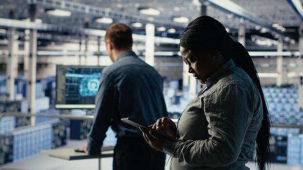 Software developers monitor server hub activity using tablet. Data center employees doing brainstorming, reviewing equipment tests results on devices to ensure systems are working correctly, camera B photo