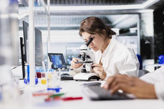 Cytotechnologist adjusting microscope, zooming on cellular structures in lab workspace. Molecular biologist observes specimens through magnification tool at laboratory bench workstation photo