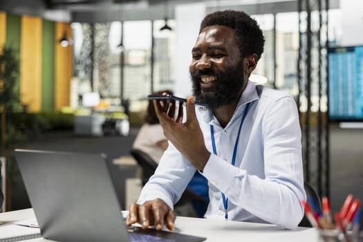 Cheerful businessman sending voice message on mobile device and using laptop. Happy male employee in office discussing ideas with coworker on phone call, solving business problems. photo