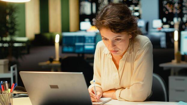 Employee works after hours organizing and scheduling tasks for a corporate project. Surrounded by files, she focuses on budgeting and objectives to achieve company targets at night. Camera B. photo