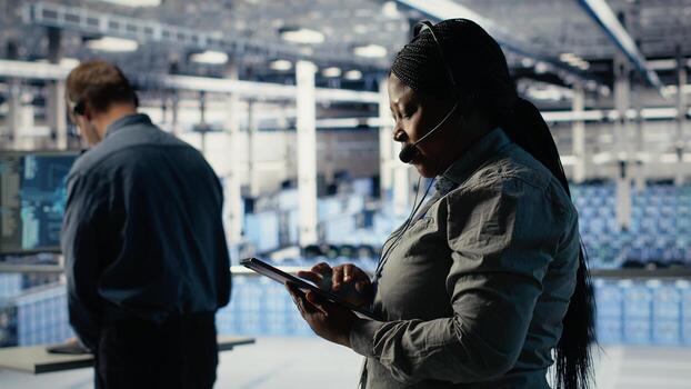 Server hub tech support expert guides enterprise clients through setting up secure company data backups. African american woman in data center helps users fix system errors on servers, camera B photo