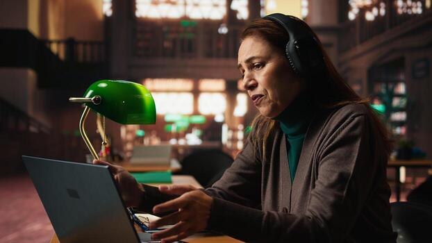 Female student participates in online class via laptop in a campus library, engaged in conference lecture while researching for a thesis and studying for academic qualifications. photo