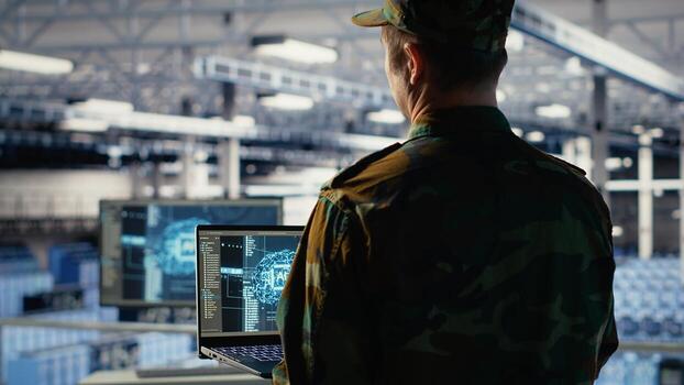 Army programmer in high tech server hub uses AI application on laptop to review reconnaissance information. Data center IT expert wearing military uniform uses AI on notebook to plan mission, camera B photo