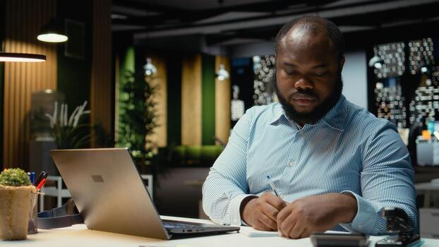 Black guy documenting essential ideas on paper and reading on laptop, planning and scheduling tasks at night. Notetaking with his handwriting for a presentation that reflects objectives. photo